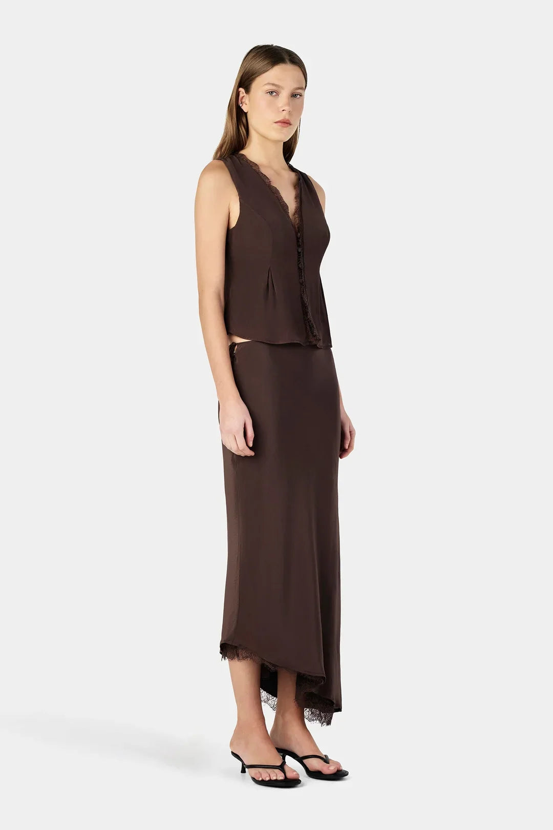 A model wearing a chocolate brown midi skirt with a flared shape and asymmetrical lace-trimmed hem, styled with a matching sleeveless top and black strappy heels, shown from a three-quarter front angle.