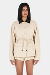 Model wearing the beige Idris Jacket with matching shorts and black heels, showing the contrast black collar, button front, patch pockets and cinched waist, front view

