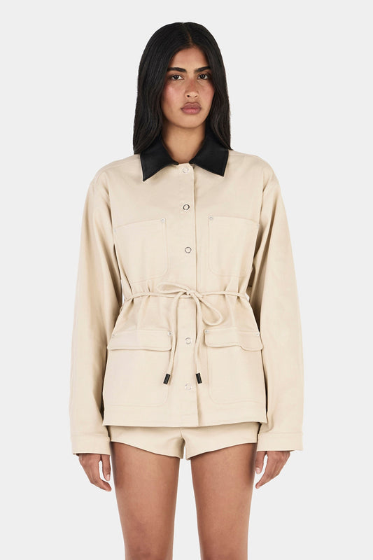 Model wearing the beige Idris Jacket with matching shorts and black heels, showing the contrast black collar, button front, patch pockets and cinched waist, front view
