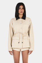 Model wearing the beige Idris Jacket with matching shorts and black heels, showing the contrast black collar, button front, patch pockets and cinched waist, front view
