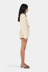 Model wearing the beige jacket with matching shorts and black heels, showing the contrast collar, relaxed sleeves and cinched waist, full-length side view
