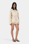 Model wearing the beige jacket with matching shorts, showing the oversized shape, button front, patch pockets and waist tie, full-length angled front view
