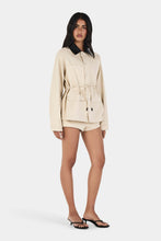 Model wearing the beige jacket with matching shorts, showing the oversized shape, button front, patch pockets and waist tie, full-length angled front view
