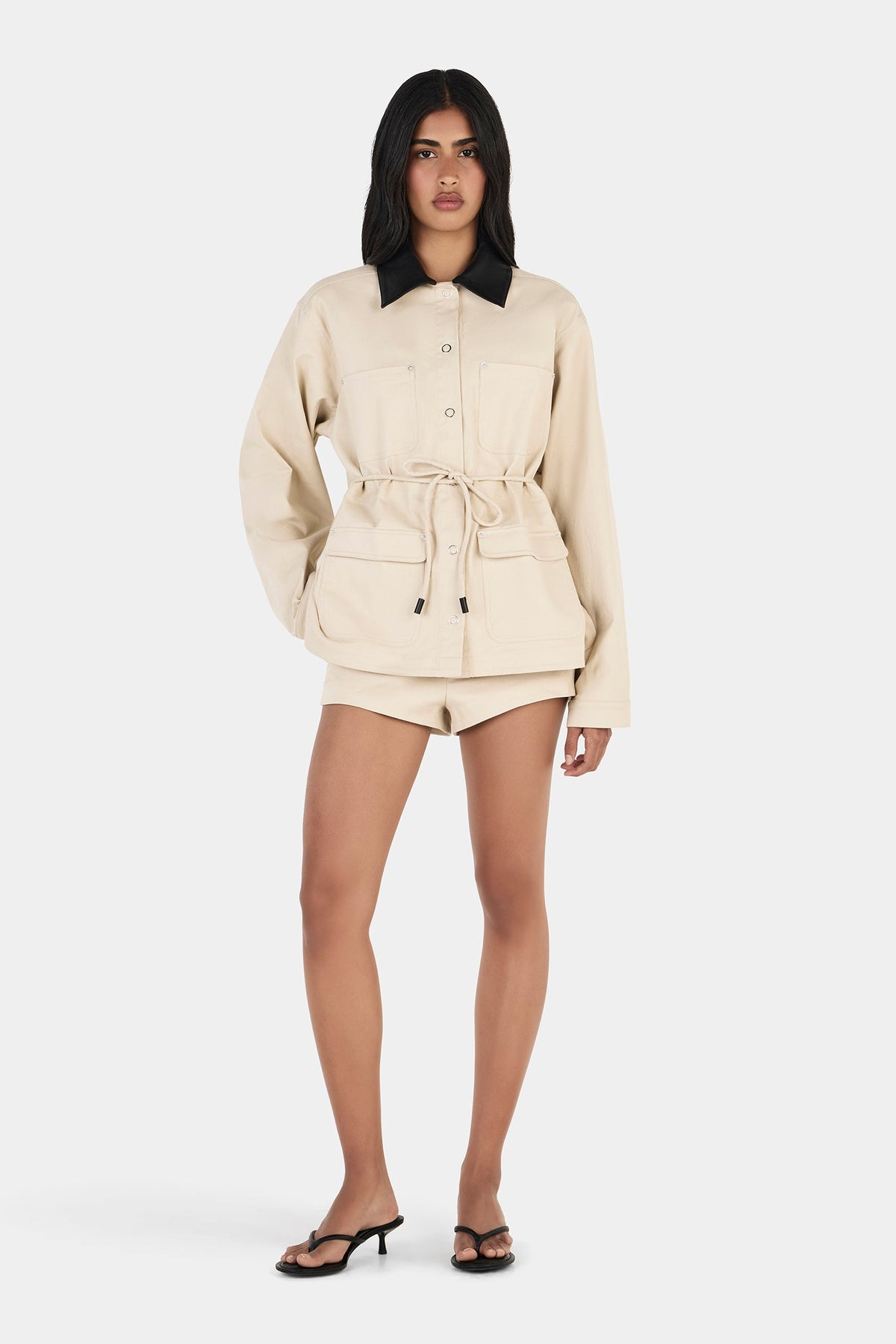 Model wearing the beige jacket with one hand on the hip, showing the oversized silhouette, patch pockets and adjustable waist tie, full-length front view
