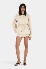 Model wearing the beige jacket with one hand on the hip, showing the oversized silhouette, patch pockets and adjustable waist tie, full-length front view
