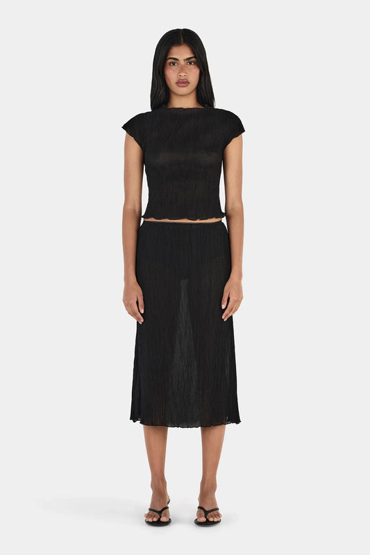 Model wearing a black midi skirt with a crinkled texture, elasticated waistband, straight silhouette, semi-sheer appearance, and slightly uneven hemline, paired with a black short-sleeve top and black heeled sandals.