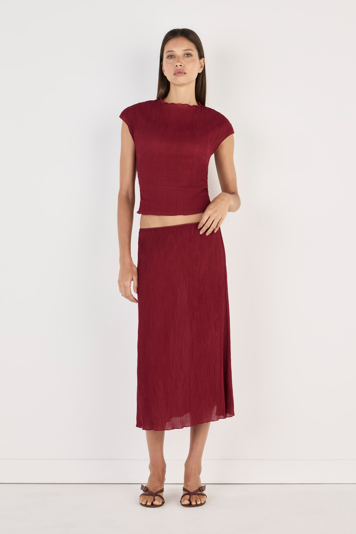 Model wearing the crimson Midi Skirt with a matching cropped top and strappy heels, showing the clean waistband, flowing midi length, lightweight crinkled texture and softly draped silhouette in a full-length front view.