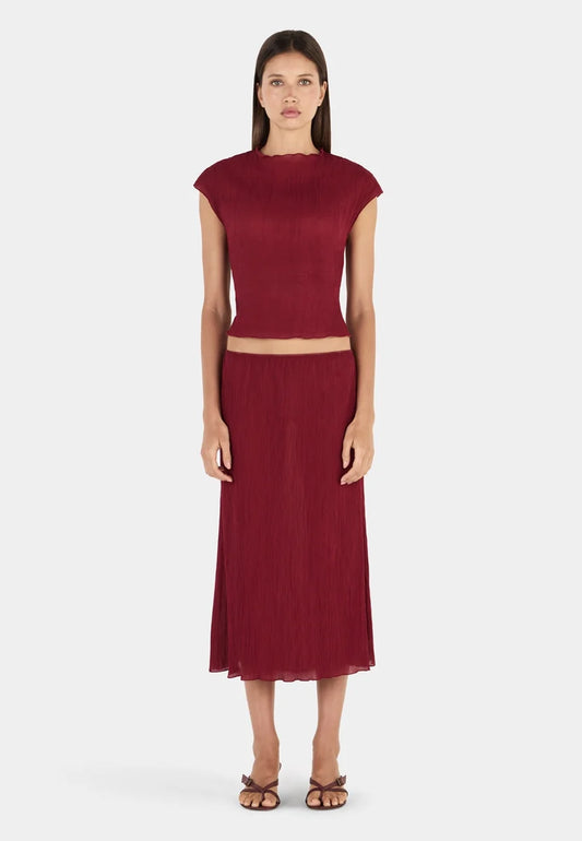 Model wearing a deep crimson midi skirt with a gathered elasticated waistband, fine crinkled texture, straight silhouette, semi-sheer appearance, and softly uneven hemline, paired with a matching short-sleeve top and brown strappy heeled sandals.
