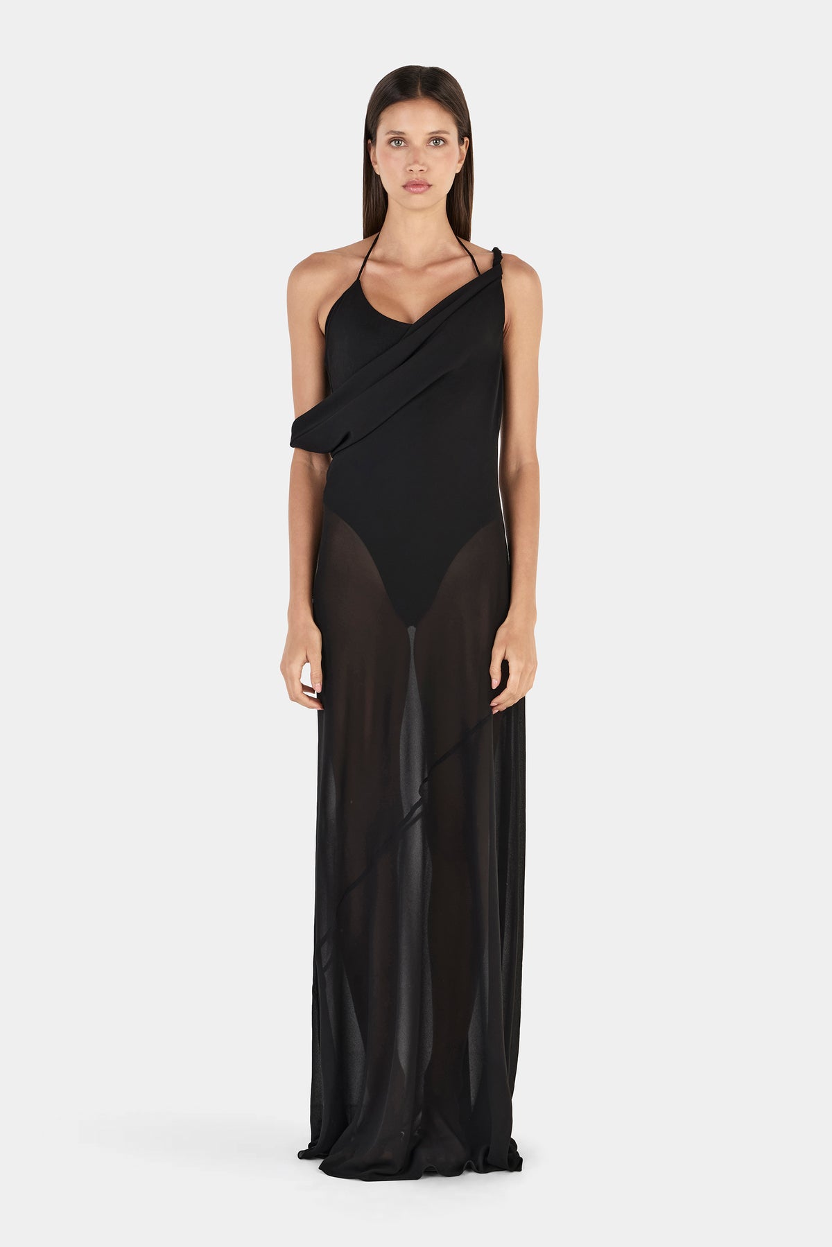 A front view of a model wearing a black maxi dress with an asymmetrical draped neckline, thin halter straps, a fitted bodysuit-style base and a sheer floor-length overlay.