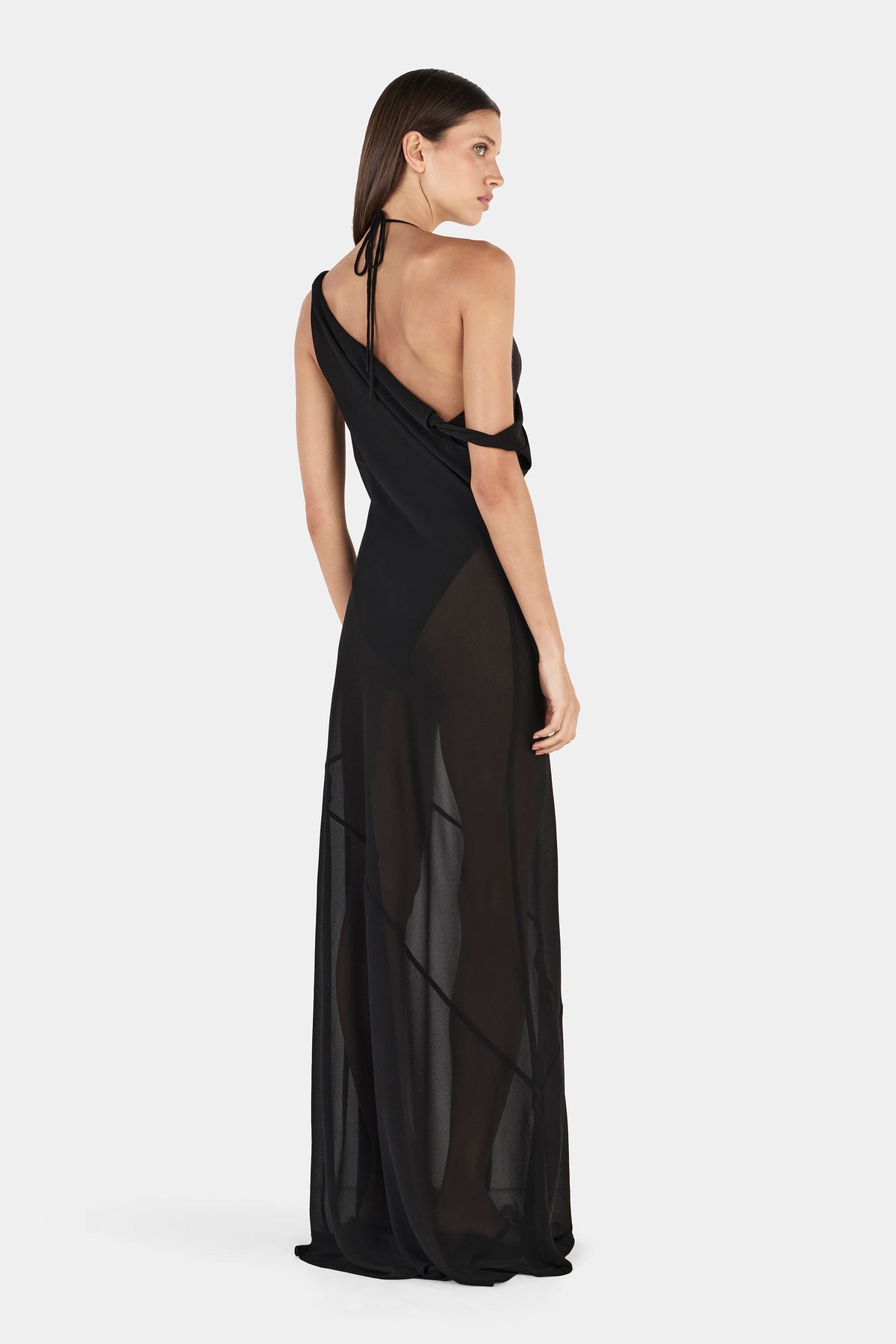 A rear three-quarter view showing the black dress's open back, tied halter straps, asymmetrical upper design and sheer flowing skirt.