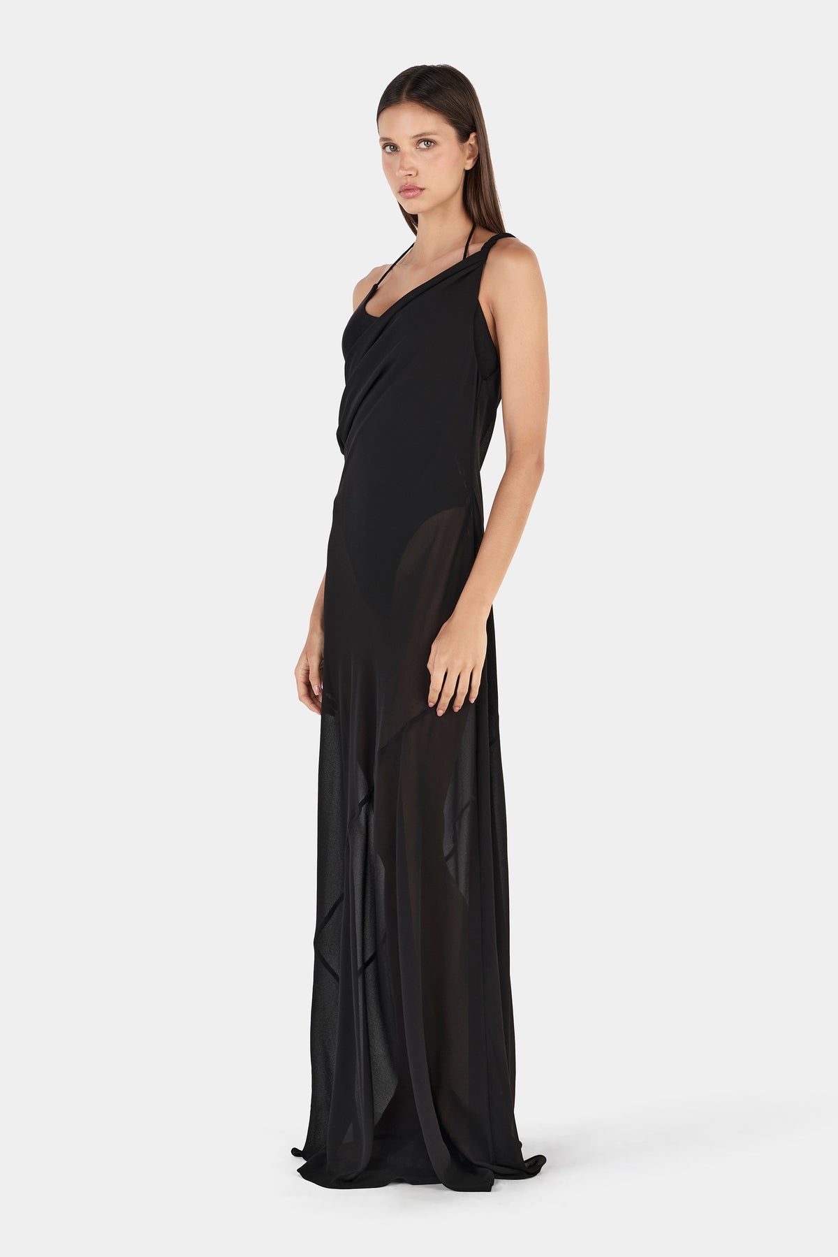 A front three-quarter view of a black maxi dress featuring an asymmetrical neckline, a fitted bodice and a sheer overlay extending to the floor.