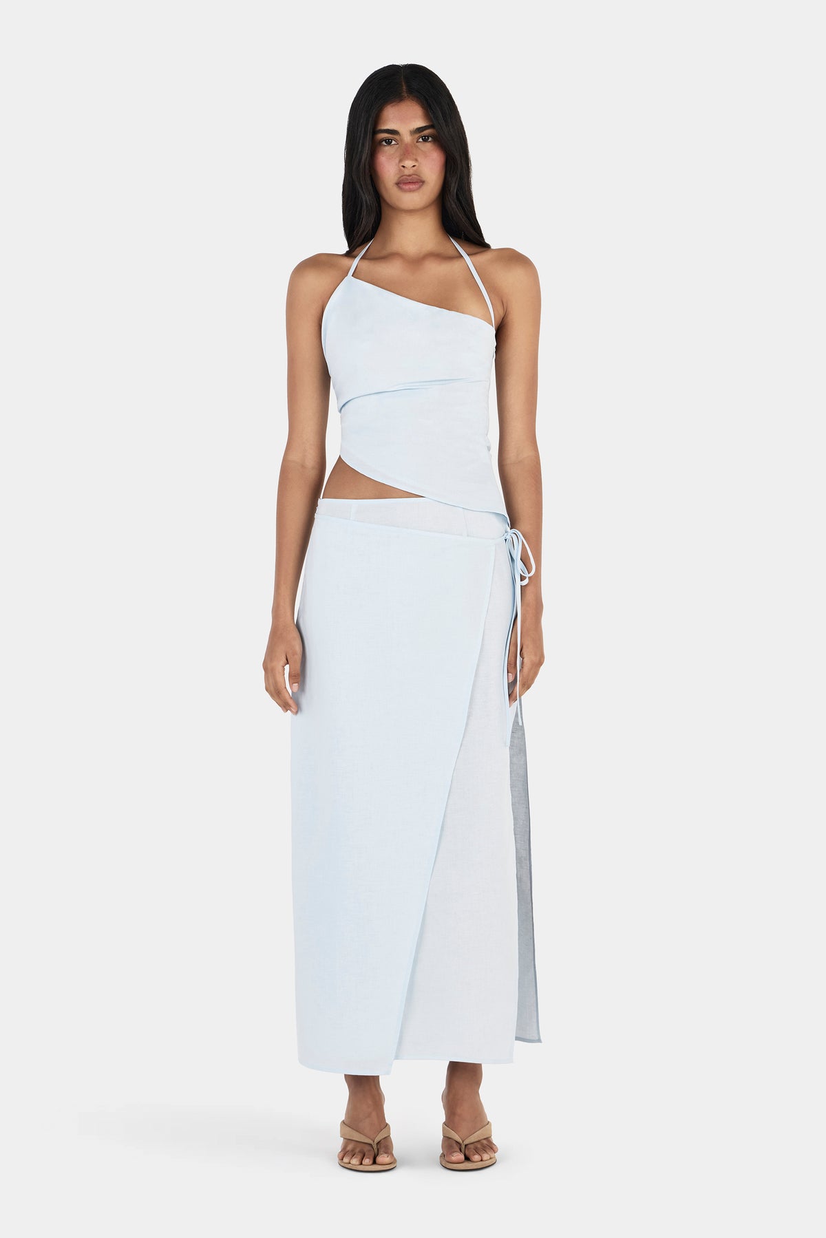 A model wearing a cornflower blue maxi skirt with a straight silhouette, overlapping wrap-style front panel and adjustable side tie at the waist, paired with a matching asymmetrical halter top and beige sandals.