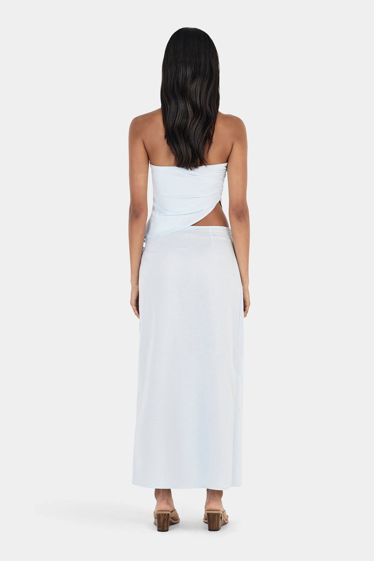 A rear view of a model wearing a cornflower blue maxi skirt with a smooth waistband, clean back panel and full-length straight silhouette, paired with a matching light blue top and brown block-heeled sandals.
