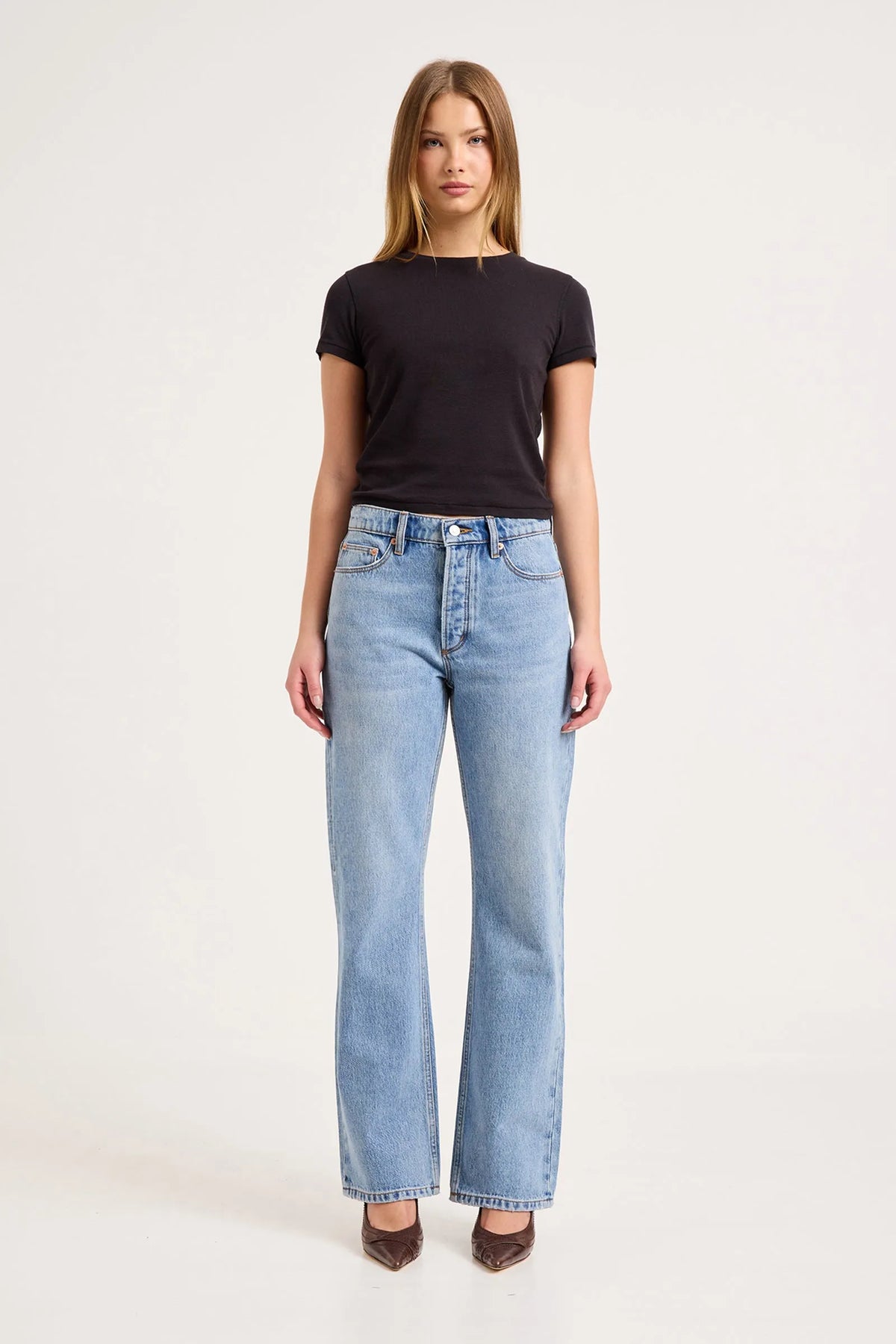 Model wearing the Surrender mid-blue boyfriend jeans with a fitted black tee and brown pointed heels, showing the mid-rise waist, relaxed fit and straight full-length legs in a front view.