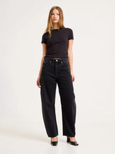 Model wearing the washed black jeans with a fitted black tee and heeled shoes, showing the relaxed seat, curved side panels and tapered barrel-leg silhouette in an angled front view.