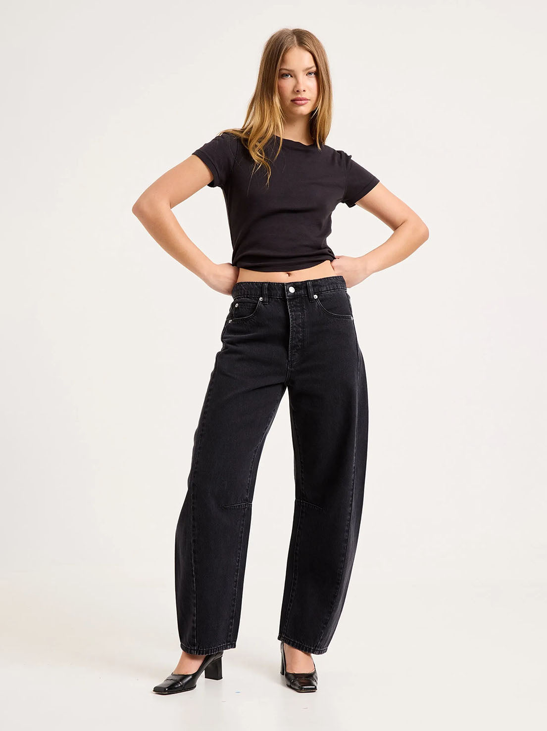 Model wearing the Mercer Black jeans with a cropped black tee and heels, showing the silver button, front pockets, side panels and curved barrel-leg shape in a styled front view.