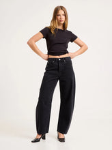 Model wearing the Mercer Black jeans with a cropped black tee and heels, showing the silver button, front pockets, side panels and curved barrel-leg shape in a styled front view.