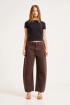 A model wears chocolate-brown barrel-leg pants with a black fitted short-sleeve top and tan sandals, showing the full-length front view, mid-rise waist and curved leg shape.
