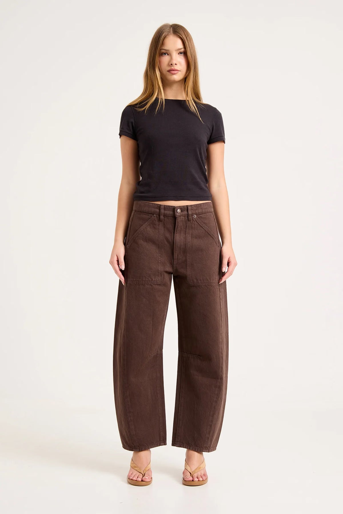 A model wears chocolate-brown barrel-leg pants with a black fitted short-sleeve top and tan sandals, showing the full-length front view, mid-rise waist and curved leg shape.