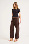 A model poses at a three-quarter angle in chocolate-brown barrel-leg pants, a fitted black short-sleeve top and tan wedge sandals, revealing the mid-rise waist, roomy leg shape, curved outer seams and utility-inspired panel details against a plain studio background.