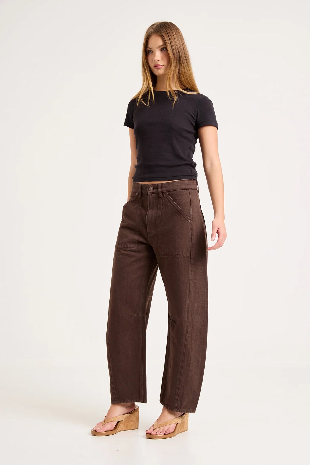 A model poses at a three-quarter angle in chocolate-brown barrel-leg pants, a fitted black short-sleeve top and tan wedge sandals, revealing the mid-rise waist, roomy leg shape, curved outer seams and utility-inspired panel details against a plain studio background.