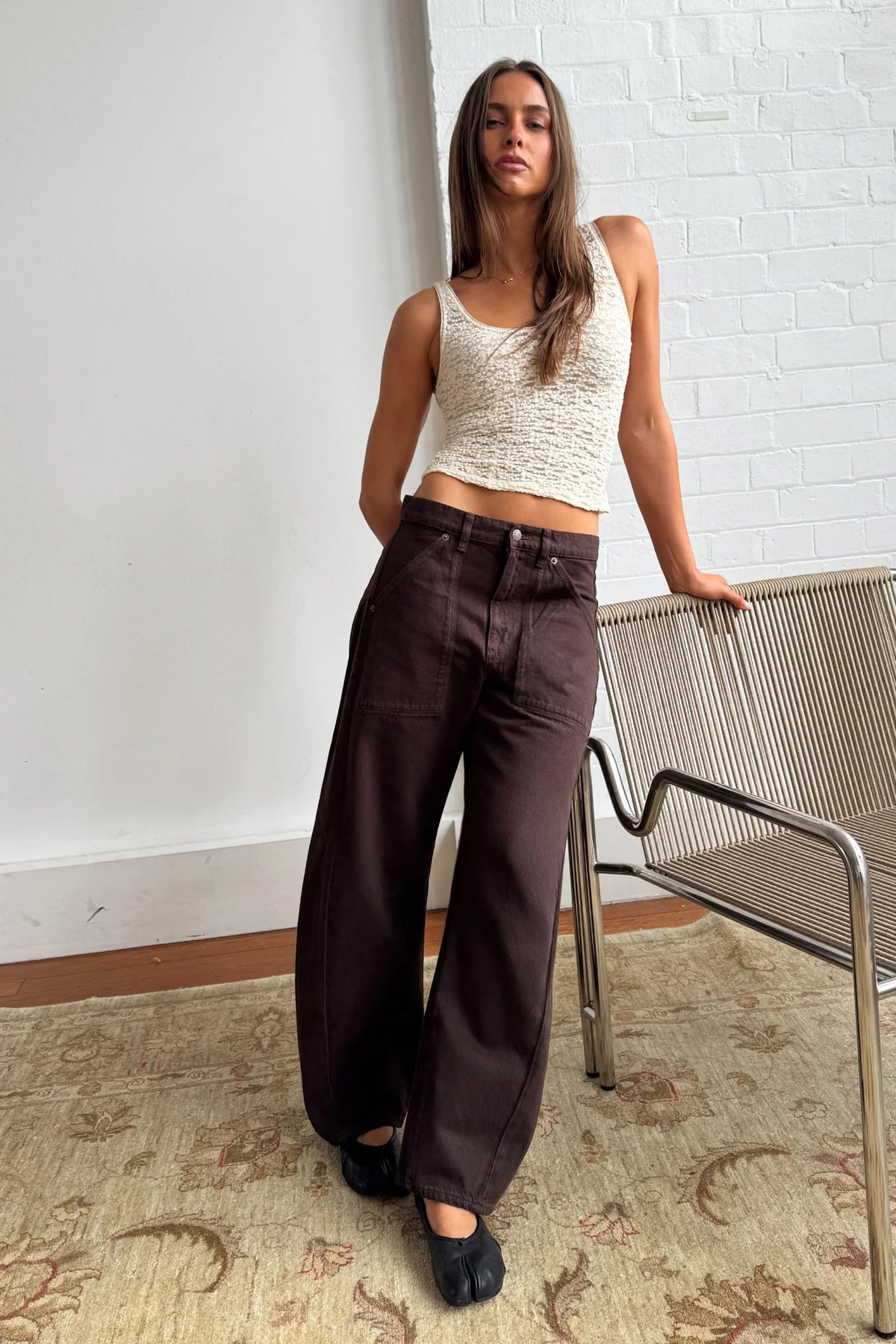 A model wears chocolate-brown barrel-leg pants with a cream textured sleeveless crop top and black ballet flats, leaning one hand against a metal-framed woven chair in a bright interior with white brick walls, a patterned rug and wooden flooring. The pose highlights the pants' relaxed volume, long curved legs and deep brown colour.