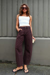 A model with curly brown hair and dark sunglasses wears chocolate-brown barrel-leg pants with a white cropped camisole and black flip-flops, posing outdoors against a grey vertical-panel wall. One hand is tucked into a front pocket while the other holds a takeaway coffee cup, showing the mid-rise waist, roomy legs and casual everyday styling.