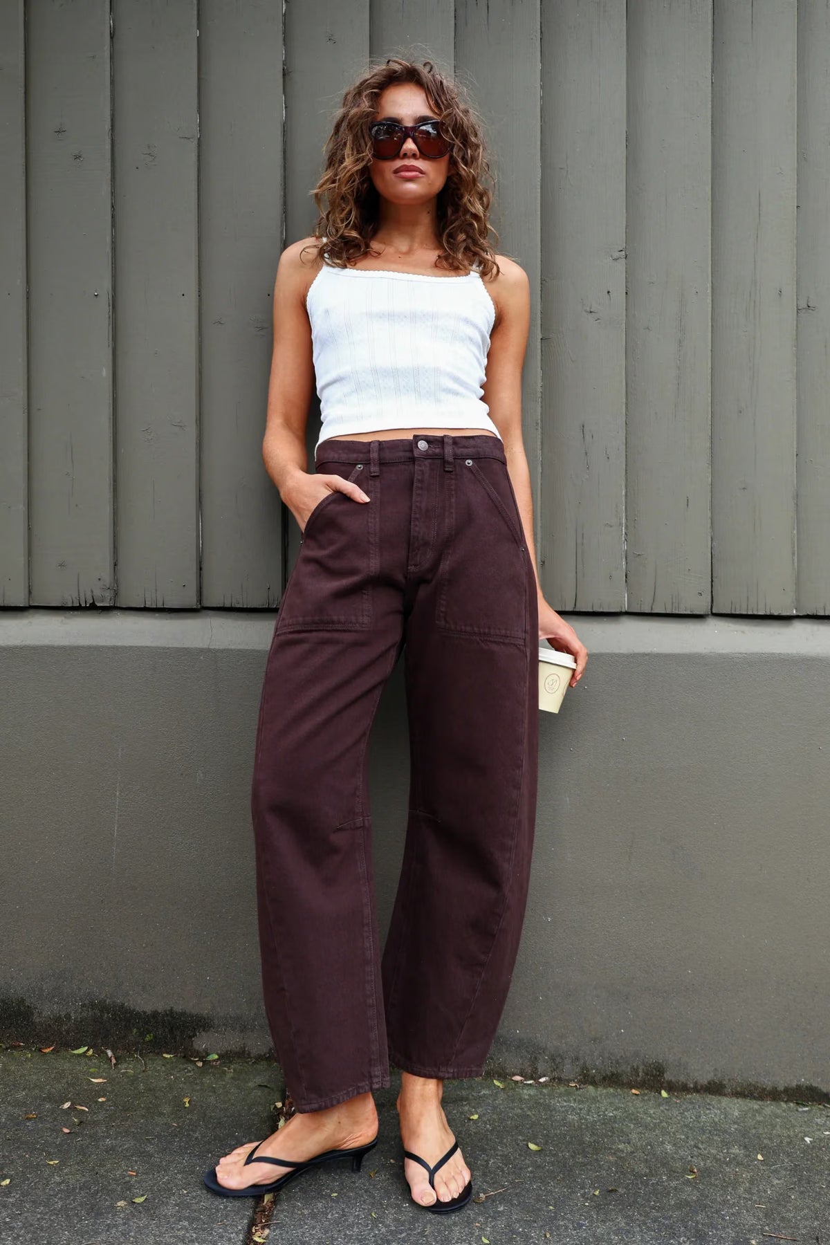 A model with curly brown hair and dark sunglasses wears chocolate-brown barrel-leg pants with a white cropped camisole and black flip-flops, posing outdoors against a grey vertical-panel wall. One hand is tucked into a front pocket while the other holds a takeaway coffee cup, showing the mid-rise waist, roomy legs and casual everyday styling.