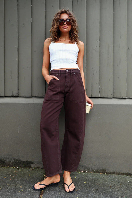 A model with curly brown hair and dark sunglasses wears chocolate-brown barrel-leg pants with a white cropped camisole and black flip-flops, posing outdoors against a grey vertical-panel wall. One hand is tucked into a front pocket while the other holds a takeaway coffee cup, showing the mid-rise waist, roomy legs and casual everyday styling.