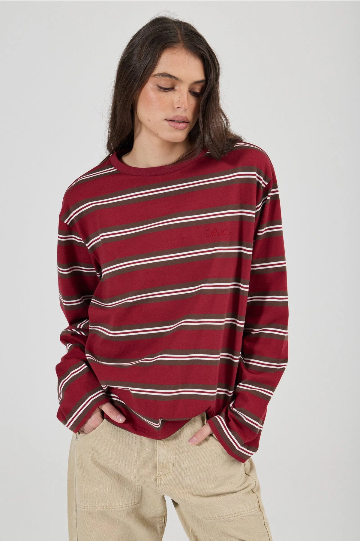Model wearing the burgundy striped long-sleeve tee with beige jeans, showing the relaxed oversized fit, crew neckline, dropped shoulders and long sleeves in an angled front view.
