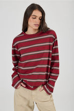 Model wearing the burgundy striped long-sleeve tee with beige jeans, showing the relaxed oversized fit, crew neckline, dropped shoulders and long sleeves in an angled front view.