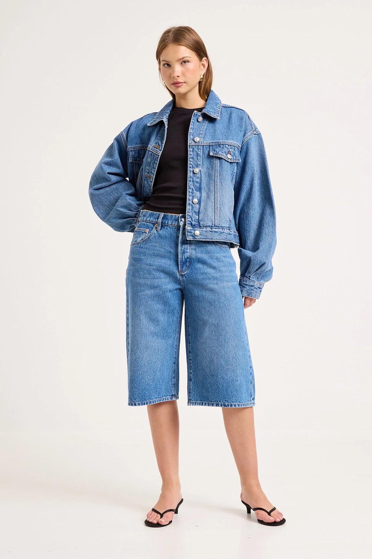 Woman wearing mid-blue denim culottes with a cropped denim jacket, black top and black heeled sandals in a studio setting.