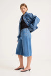 Woman wearing mid-blue denim culottes with a denim jacket and black heeled sandals, shown from a three-quarter side view.