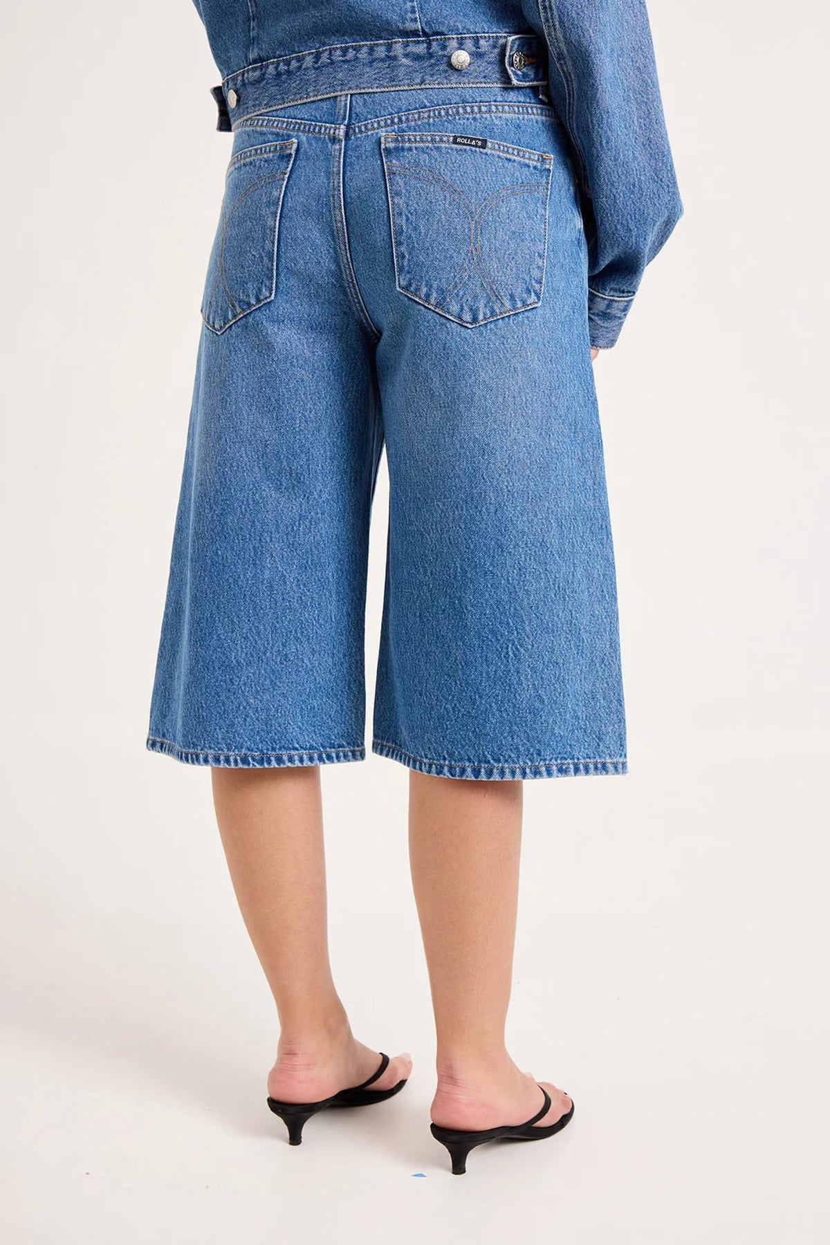 Close-up back view of mid-blue denim culottes showing the two square back pockets, contrast stitching and below-knee length.