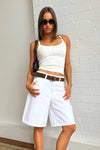 Woman wearing white knee-length denim shorts with a fitted white top and brown belt against a white brick wall.