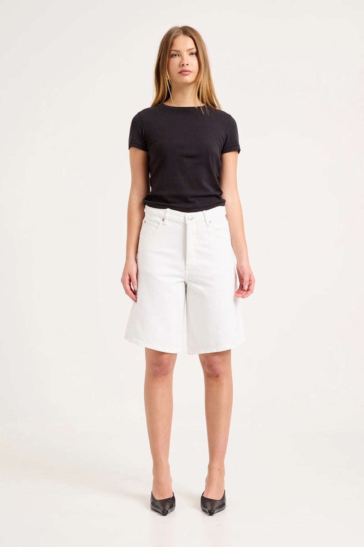 Woman wearing white knee-length denim shorts with a black fitted T-shirt and black pointed shoes in a studio setting.