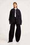 Model wearing the Mercer Black denim chore jacket over a white tee with black wide-leg trousers and pointed shoes, showing the contrast collar, silver button front, chest pocket, lower patch pockets and relaxed boxy fit.