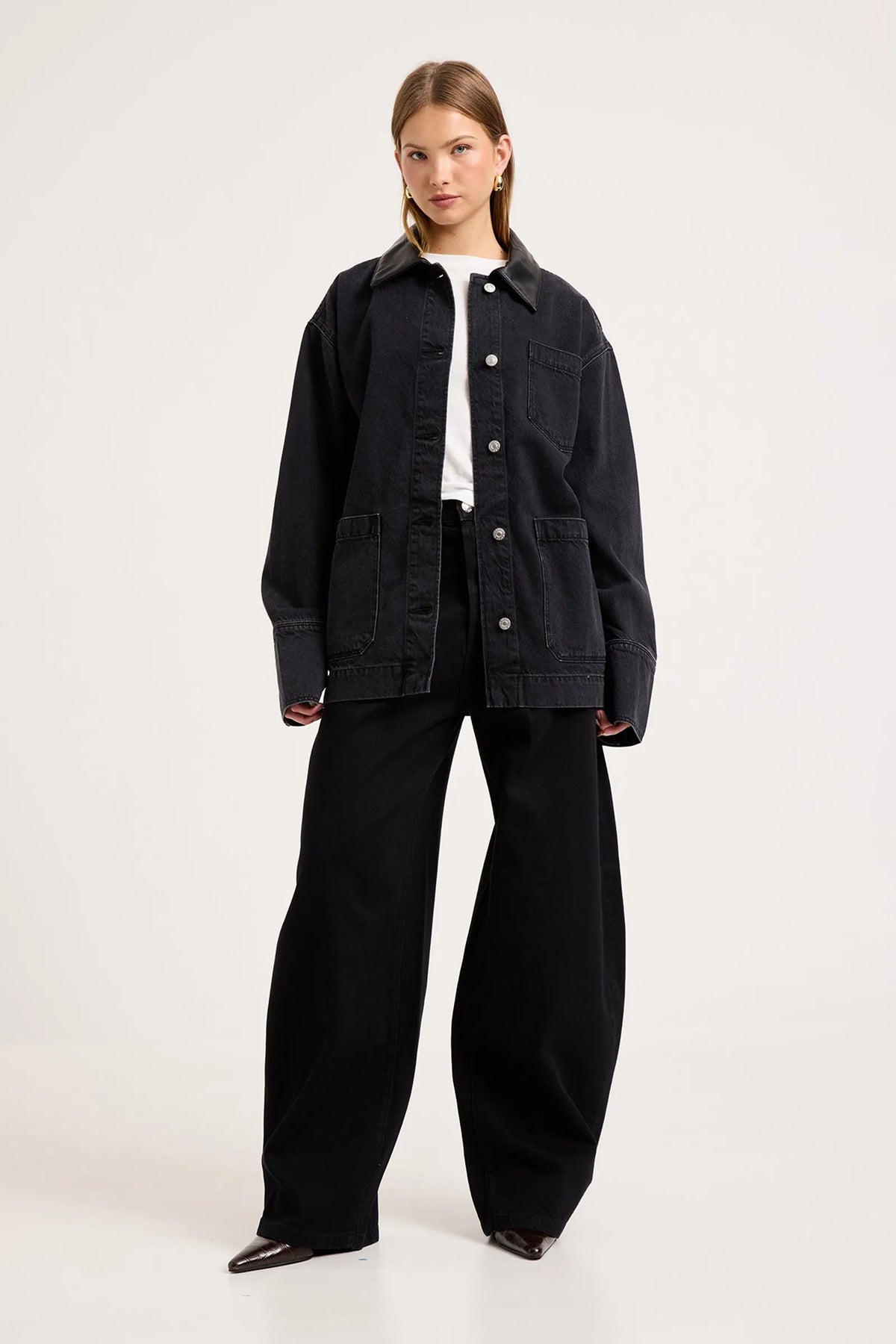 Model wearing the Mercer Black denim chore jacket over a white tee with black wide-leg trousers and pointed shoes, showing the contrast collar, silver button front, chest pocket, lower patch pockets and relaxed boxy fit.