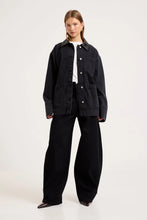 Model wearing the Mercer Black denim chore jacket over a white tee with black wide-leg trousers and pointed shoes, showing the contrast collar, silver button front, chest pocket, lower patch pockets and relaxed boxy fit.