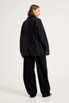 Model wearing the black denim chore jacket with matching wide-leg trousers, showing the dropped shoulders, straight back, long sleeves and oversized workwear-inspired silhouette from the back.