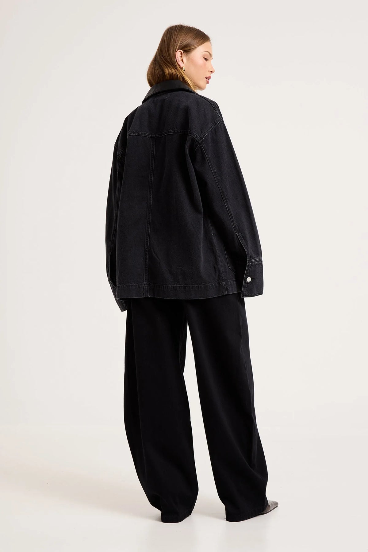 Model wearing the black denim chore jacket with matching wide-leg trousers, showing the dropped shoulders, straight back, long sleeves and oversized workwear-inspired silhouette from the back.