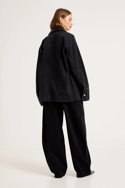 Model wearing the black denim chore jacket with matching wide-leg trousers, showing the dropped shoulders, straight back, long sleeves and oversized workwear-inspired silhouette from the back.