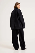 Model wearing the black denim chore jacket with matching wide-leg trousers, showing the dropped shoulders, straight back, long sleeves and oversized workwear-inspired silhouette from the back.
