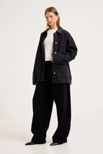 Model wearing the black denim chore jacket open over a white tee with black wide-leg trousers, showing the traditional collar, silver hardware, chest pocket, lower patch pockets and relaxed longline shape.