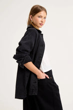 Close-up side view of the black denim chore jacket worn over a white tee, showing the contrast collar, dropped shoulder, rolled sleeve, washed denim texture and relaxed oversized fit.