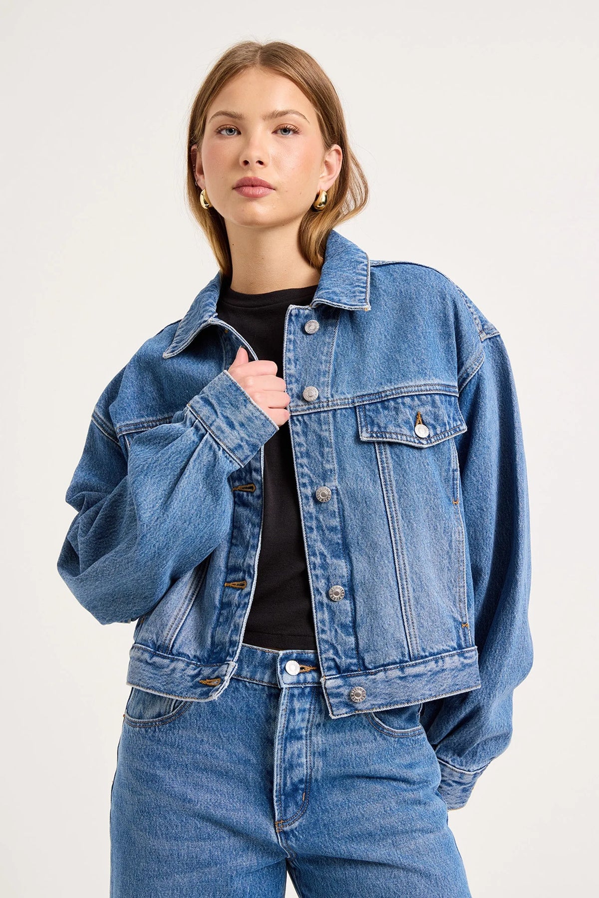 Model wearing the blue denim swing jacket over a black fitted top with matching jeans, showing the relaxed silhouette, pointed collar, flap chest pockets and silver button fastening, close-up front view
