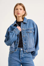 Model wearing the blue denim swing jacket over a black fitted top with matching jeans, showing the relaxed silhouette, pointed collar, flap chest pockets and silver button fastening, close-up front view
