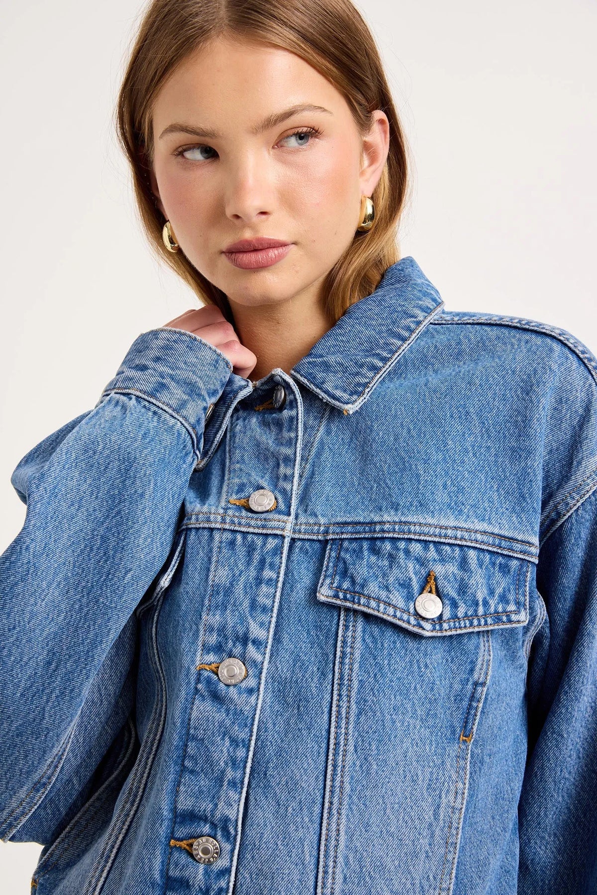 Close-up of a model wearing the Soho Vintage blue denim swing jacket, showing the pointed collar, silver button front, flap chest pocket, gold topstitching, dropped shoulders and oversized sleeve detail.