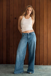 Model wearing the Soho Vintage mid-blue wide-leg jeans with a fitted white tank, showing the mid-rise waist, relaxed seat and full-length loose-leg silhouette in a lifestyle front view.