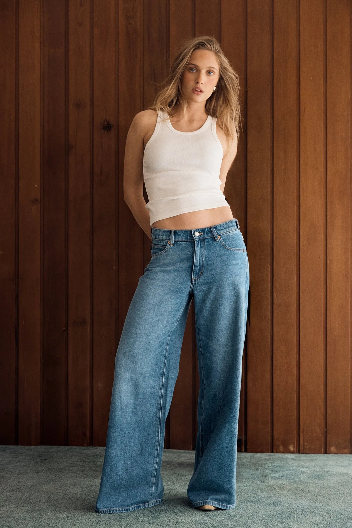 Model wearing the Soho Vintage mid-blue wide-leg jeans with a fitted white tank, showing the mid-rise waist, relaxed seat and full-length loose-leg silhouette in a lifestyle front view.